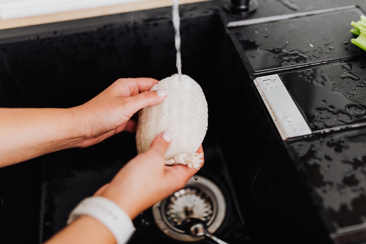 Close-up of hands cleaning a sponge under tap water in a black sink, emphasizing hygiene and kitchen cleanliness.