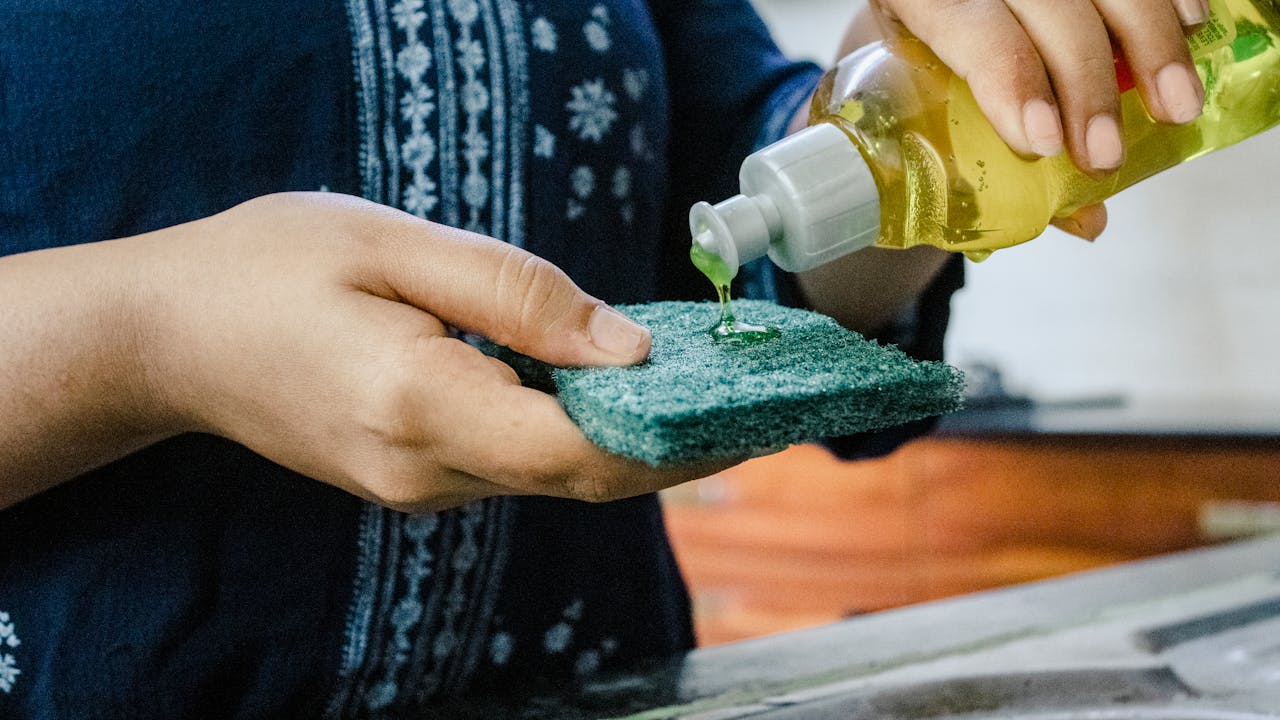 Person pouring green dishwashing soap onto a scrub pad in a kitchen setting.